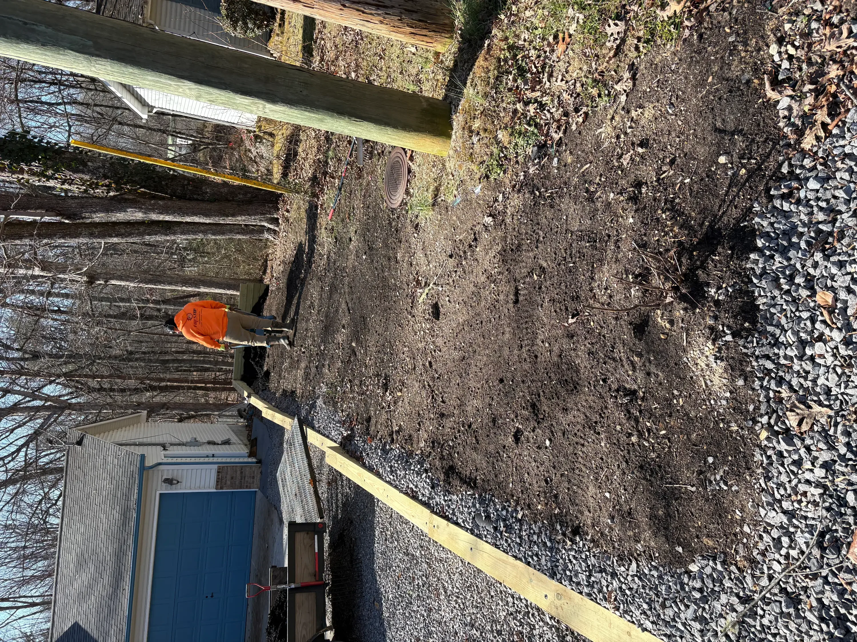 Worker in orange safety gear pressure washing moss-covered concrete driveway and foundation